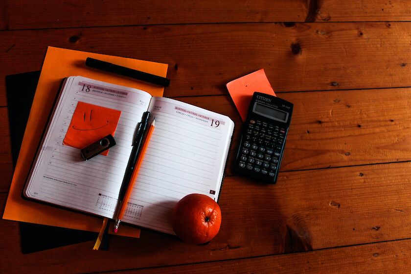 Holztisch im Hintergrund, darauf aufgeklapptes Notizbuch mit Kugelschreiber, Taschenrechner und Apfel